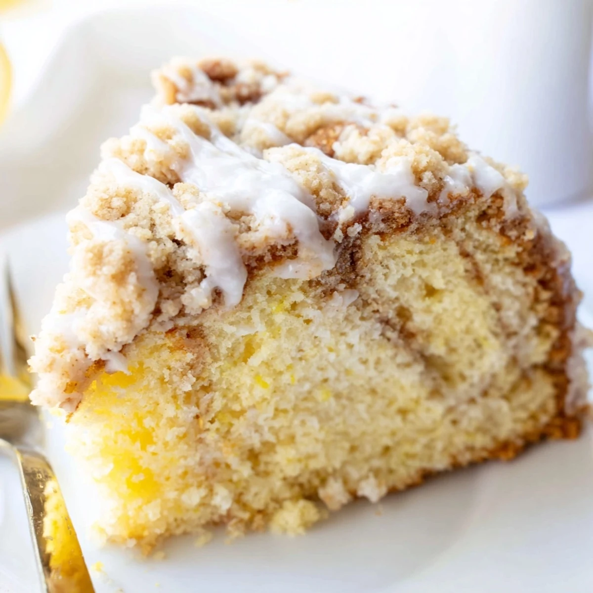 A slice of Lemon Coffee Cake with Crumble is drizzled with sweet lemon glaze on a white plate.