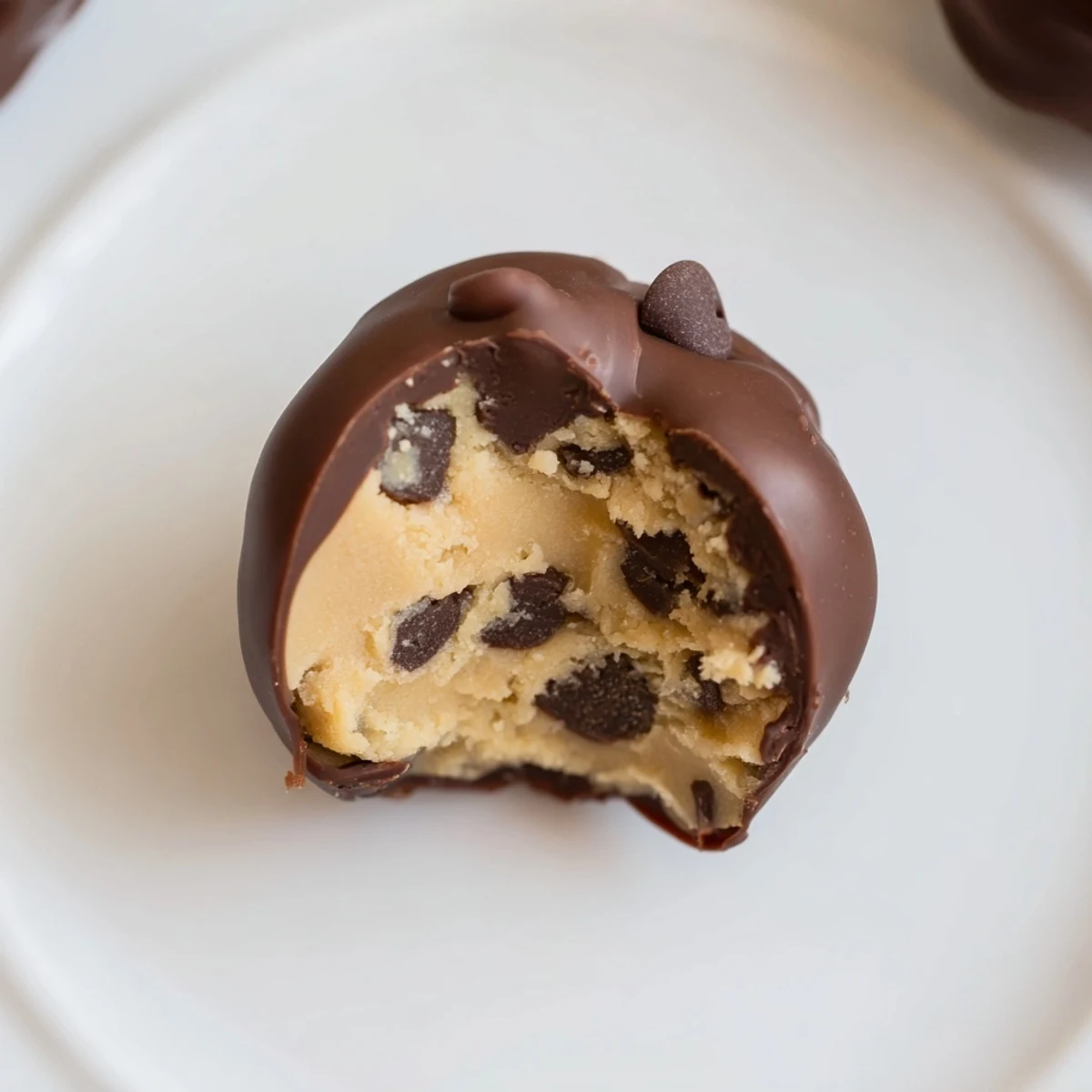 Four glossy Chocolate Chip Cookie Dough Truffles rest on a chilled tray, their smooth dark chocolate shells gleaming under soft kitchen light.
