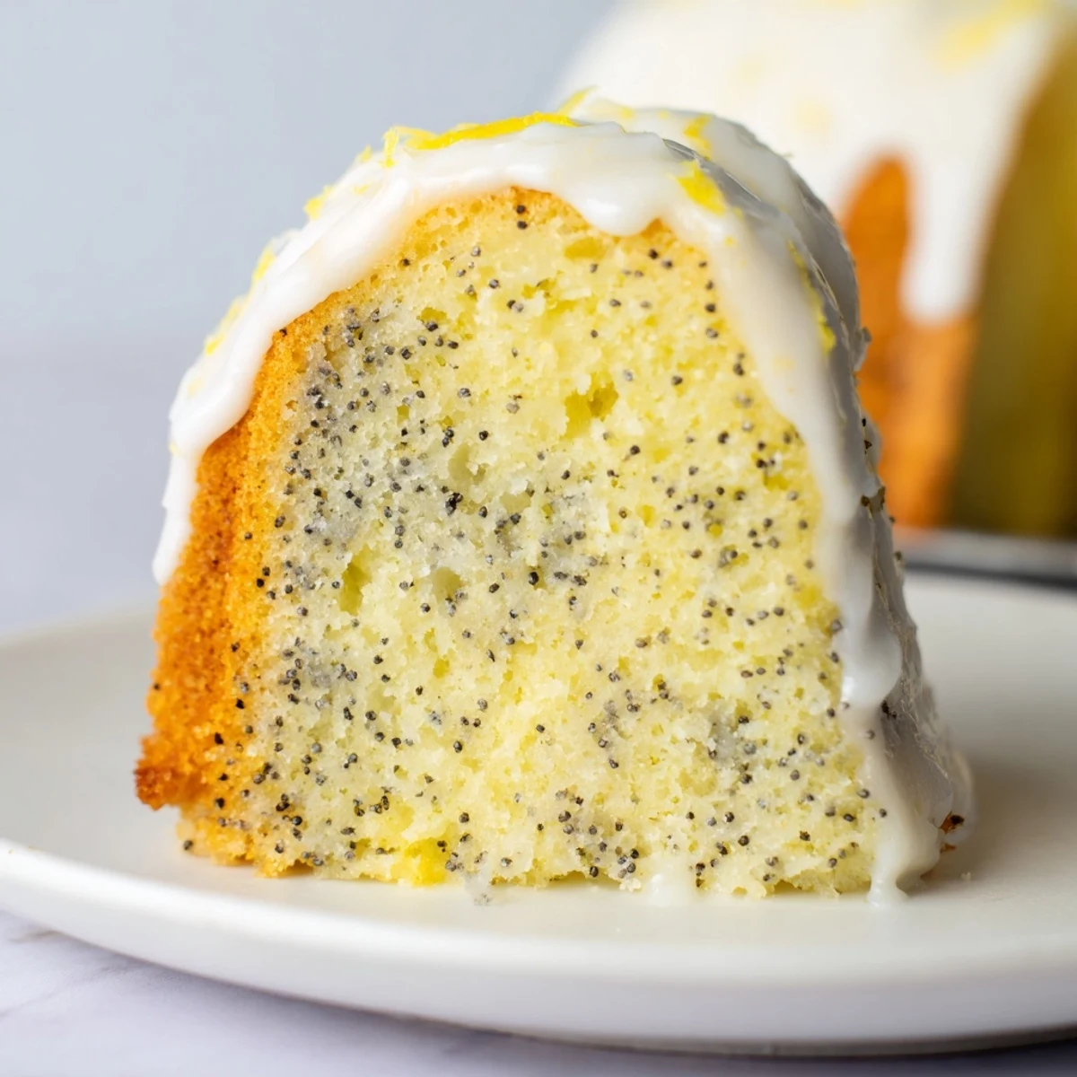 A close-up of a Lemon Poppy Seed Bundt Cake with Icing drizzled over the moist ridges, served on a white cake stand.