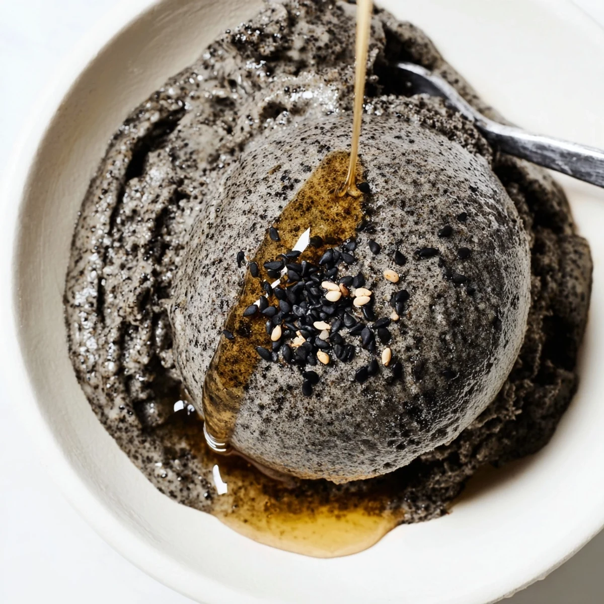 Creamy toasted black sesame ice cream served in a bowl, topped with a delicate honey drizzle and sesame seeds for a nutty, dessert-forward presentation.