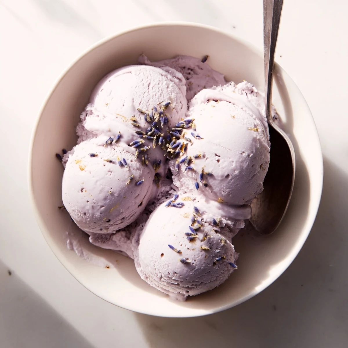 A close-up of homemade Lavender Ice Cream scoops in a waffle cone, with a swirl of creamy purple hue and a sprig of fresh lavender on top for garnish.