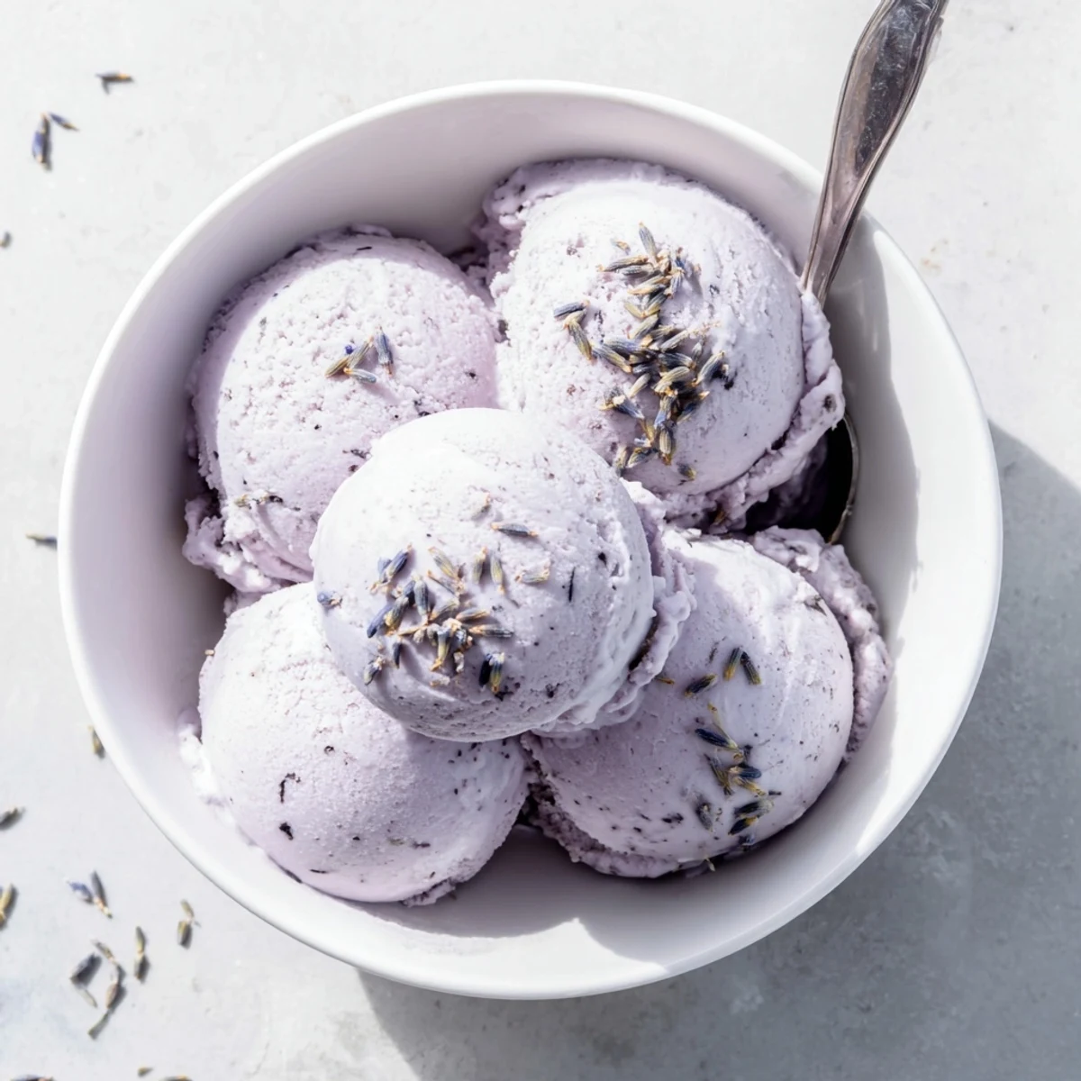 A scoop of Lavender Ice Cream melting slightly in a glass bowl, revealing smooth texture next to shortbread cookies and fresh berries on a rustic table.