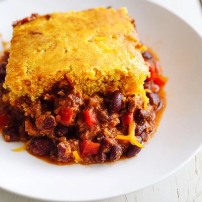 Freshly baked Beef Chili with Cornbread Topping casserole slice on a plate, topped with a dollop of sour cream and chopped green onions.